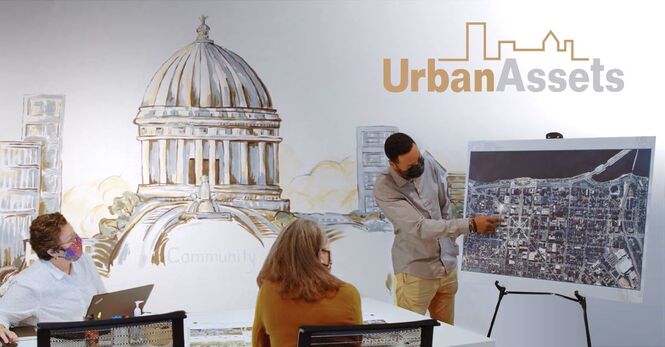 A photo of three people in a conference room, looking at a map. The Urban Assets logo is in the top-right corner of the image. 
