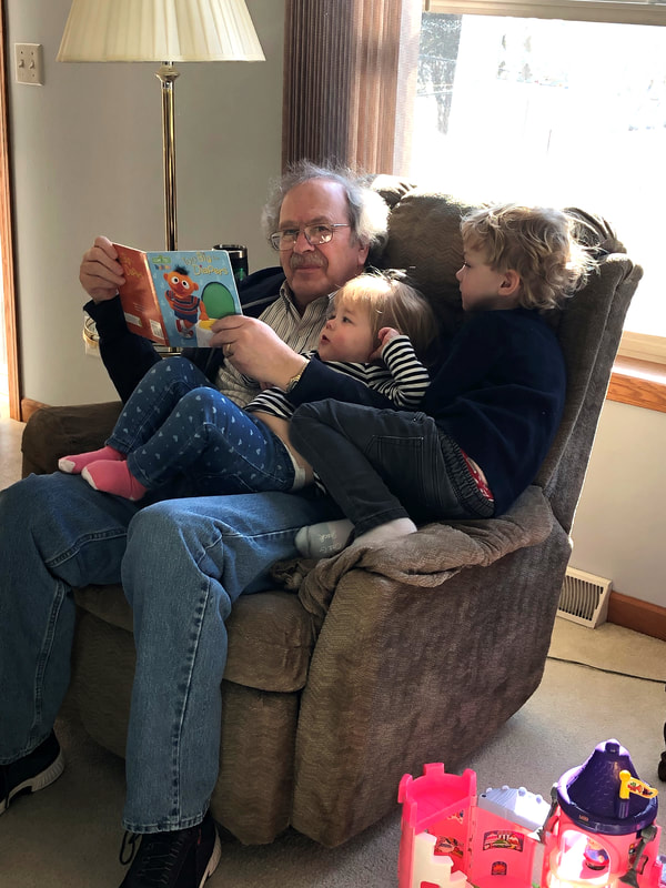 A photo of Greg sitting in an armchair and reading a book to his grandchildren.