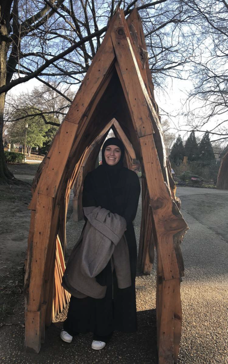 A photo of Mary outside standing beneath a wooden sculpture. 