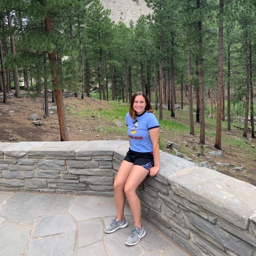 Photo of Rachel sitting on a stone half wall outdoors. A wooded area behind her.